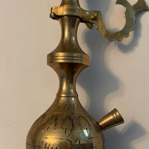 May include: A tall, polished brass vessel featuring a wide, flared top opening, a slender neck, and a bulbous base. The body is decorated with etched leaf-like patterns. A curved handle is attached to the neck, and a small, angled spout protrudes from the side of the base.