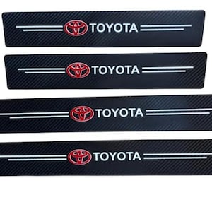 Puede incluir: Juego de cuatro protectores de umbral de puerta de coche negros con textura de fibra de carbono. Cada pieza presenta el logotipo rojo de Toyota y el nombre de la marca en blanco. Líneas paralelas blancas acentúan el logotipo.