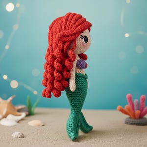 May include: Crocheted mermaid doll with red hair, a tan face, and a green tail. The doll has a purple top and is posed on a sandy surface with a blue background. The doll is approximately 10 inches tall.