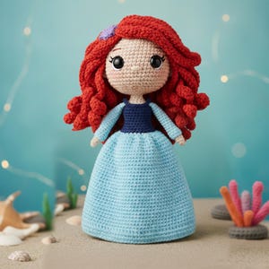 May include: A handmade crochet doll with red hair, a blue dress, and a purple hair accessory. The doll has large black eyes and a tan face. The doll is standing on a sandy surface with a blue background.