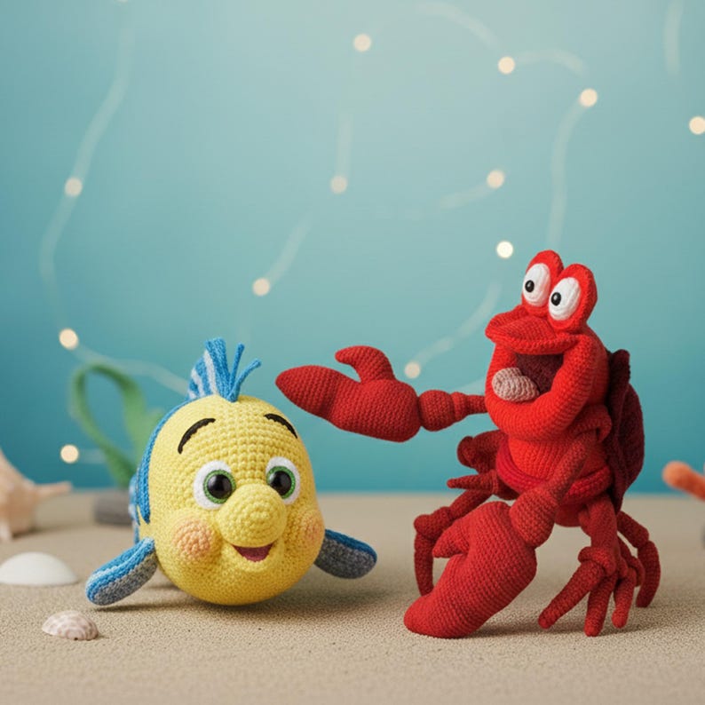May include: Crocheted Flounder and Sebastian the crab toys. Flounder is yellow with blue accents, and Sebastian is red. They are set on a sandy surface with a light blue background and string lights.