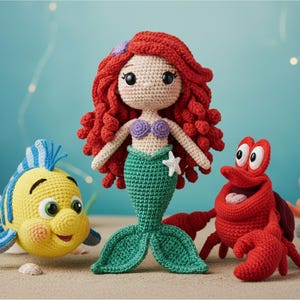 May include: Crocheted mermaid doll with red hair, a green tail, and a purple seashell top, flanked by a yellow fish and a red crab. The mermaid holds a white starfish. The toys are set on a sandy surface against a blue background.