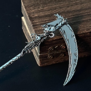 Puede incluir: Una guadaña plateada con detalles intrincados, incluyendo un cráneo y rasgos de dragón. La hoja curva tiene un borde decorativo. La guadaña se encuentra sobre un fondo oscuro y una caja de madera.