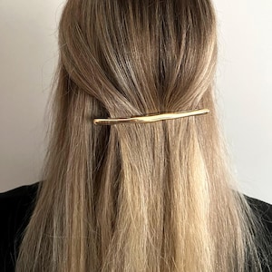 May include: A gold-colored hair barrette is positioned horizontally across the back of the head, securing a portion of blonde hair. The barrette has a textured, organic design. The hair is styled with a slight wave.