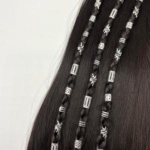 May include: Close-up of black braided hair adorned with silver beads and decorative charms. The beads are cylindrical and rectangular, with various patterns. The charms feature leaf-like designs, adding a touch of elegance to the hairstyle.
