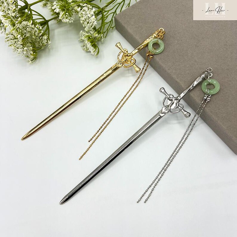 Op de afbeelding: Twee decoratieve haarstokjes in goud- en zilverkleur. Elk stokje heeft een zwaardachtig ontwerp met een decoratieve knoop en een groene cirkelvormige steen. Delicate kettingkwastjes hangen aan elk stokje en voegen elegantie toe.
