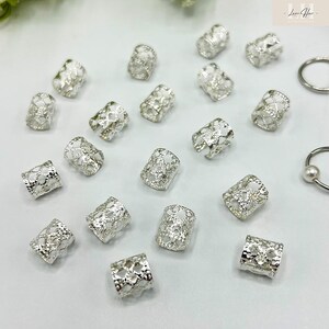 May include: A collection of silver-colored hair beads with an openwork design. The cylindrical beads are scattered across a white surface, with a few rings visible in the background. The beads have a delicate, lacy pattern.