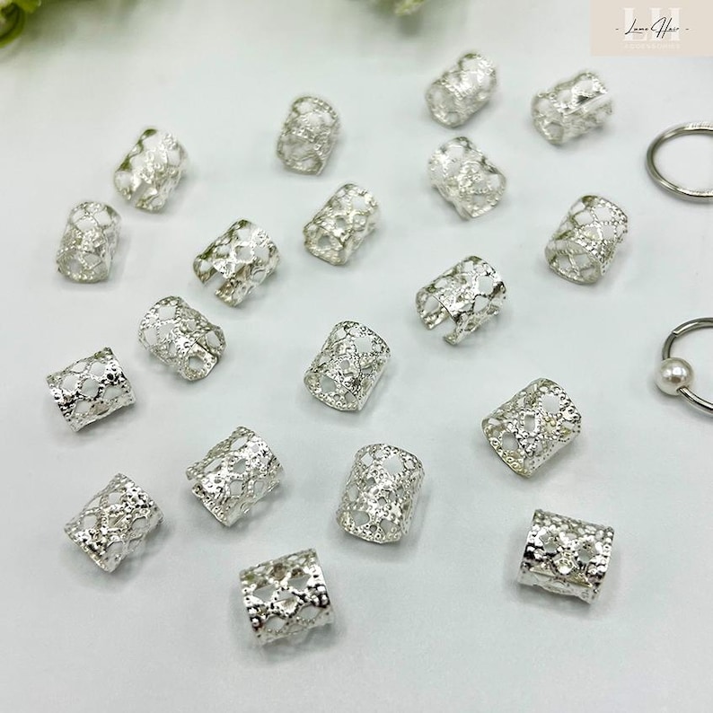 May include: A collection of silver-colored hair beads with an openwork design. The cylindrical beads are scattered across a white surface, with a few rings visible in the background. The beads have a delicate, lacy pattern.