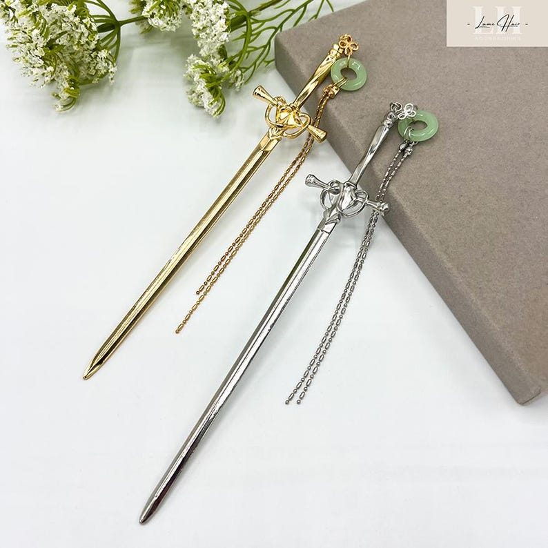 Op de afbeelding: Twee decoratieve haarspelden in de vorm van zwaarden. E&eacute;n is goudkleurig, de andere zilverkleurig. Elk heeft een ketting en een jade-kleurige ring aan de bovenkant. De achtergrond is wit met een takje witte bloemen en een grijze doos.