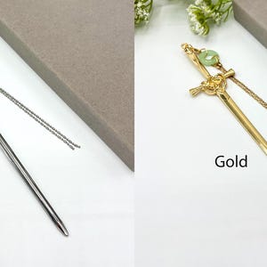 Op de afbeelding: Zilveren en goudkleurige haarstokjes met een zwaardontwerp. Elke stok heeft een decoratieve groene kraal en een bungelende ketting. De woorden "Silver" en "Gold" staan op de afbeelding.