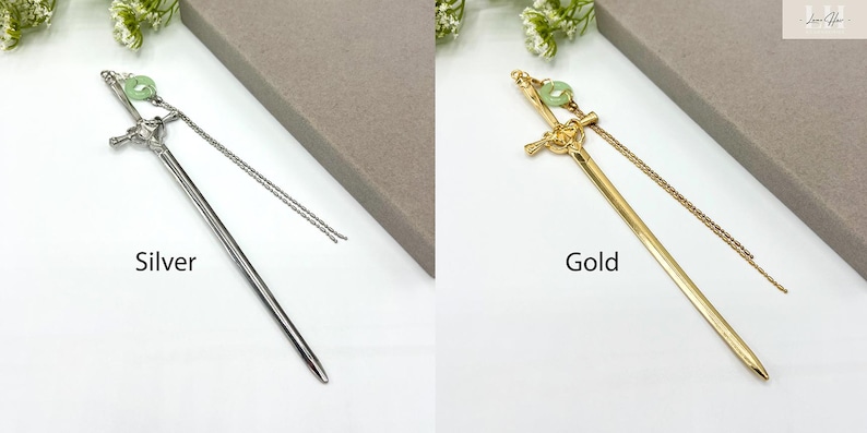 Op de afbeelding: Zilveren en goudkleurige haarstokjes met een zwaardontwerp. Elke stok heeft een decoratieve groene kraal en een bungelende ketting. De woorden "Silver" en "Gold" staan op de afbeelding.