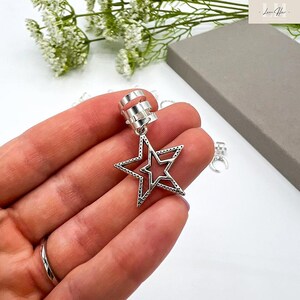 May include: Silver-colored ear cuff with a dangling star pendant. The star has a cutout design and is accented with small dots around the edges. The ear cuff has a three-band design for secure placement.