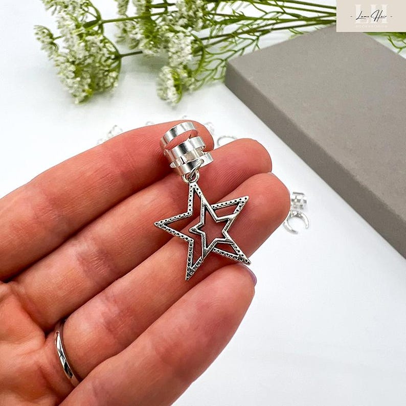 May include: Silver-colored ear cuff with a dangling star pendant. The star has a cutout design and is accented with small dots around the edges. The ear cuff has a three-band design for secure placement.