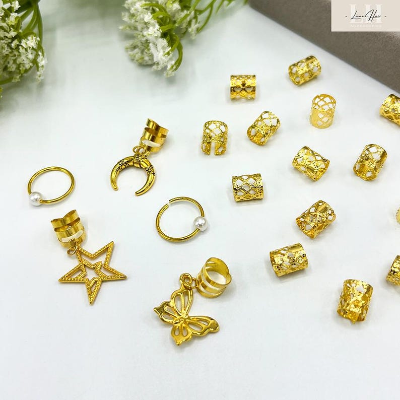 May include: Gold-colored hair jewelry, including rings with pearls, star and butterfly charms, crescent moon charms, and cylindrical beads. The accessories are scattered on a white surface, with some floral accents in the background.