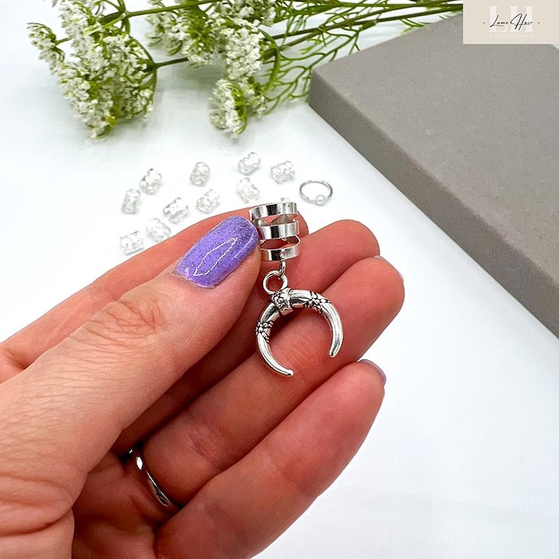 May include: Silver-toned hair cuff with a crescent moon design. The cuff features a series of stacked rings at the top, connected to a crescent moon pendant with floral detailing. The jewelry is held in a hand with a purple nail.