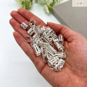 以下が含まれることがあります： 様々なサイズのビーズやリングを含む、銀色のヘアアクセサリーのコレクションが手に持たれています。アクセサリーは複雑な透かし彫りのデザインです。画像は明るく照らされており、アイテムの金属的な輝きを際立たせています。