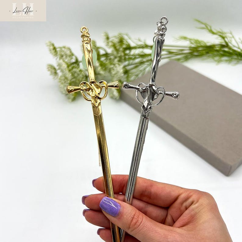 Op de afbeelding: Twee decoratieve haarstokjes in een zwaardontwerp. E&eacute;n is goudkleurig, de andere zilverkleurig, beide met een groene edelsteen aan de bovenkant. De handvatten hebben ingewikkelde hart- en kruisontwerpen. De stokjes worden tegen een witte achtergrond gehouden.