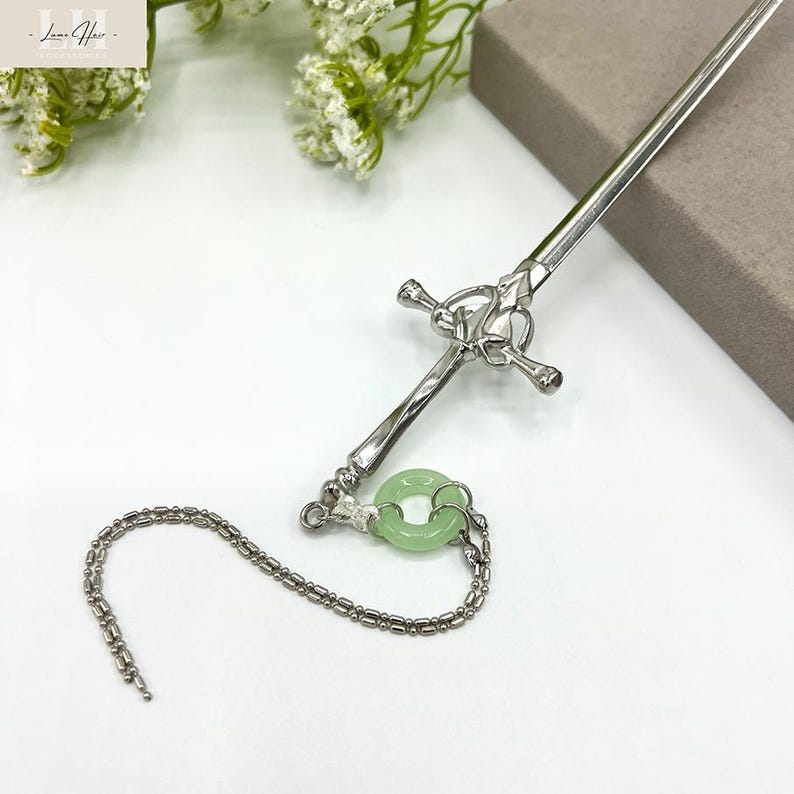 Op de afbeelding: Zilverkleurige haarspeld met een zwaardachtig ontwerp, voorzien van een decoratieve kruisbescherming en een lichtgroene jade ring hanger, bevestigd met een ketting. Het haaraccessoire staat op een witte achtergrond.