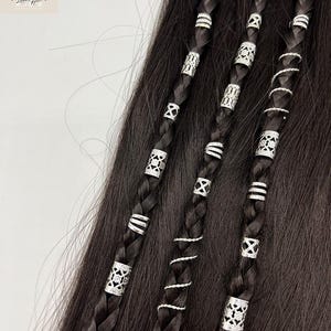 May include: Close-up of dark brown braided hair adorned with silver hair beads and spiral wraps. The beads feature various geometric designs, adding a decorative touch to the hairstyle. The hair is styled in multiple braids.