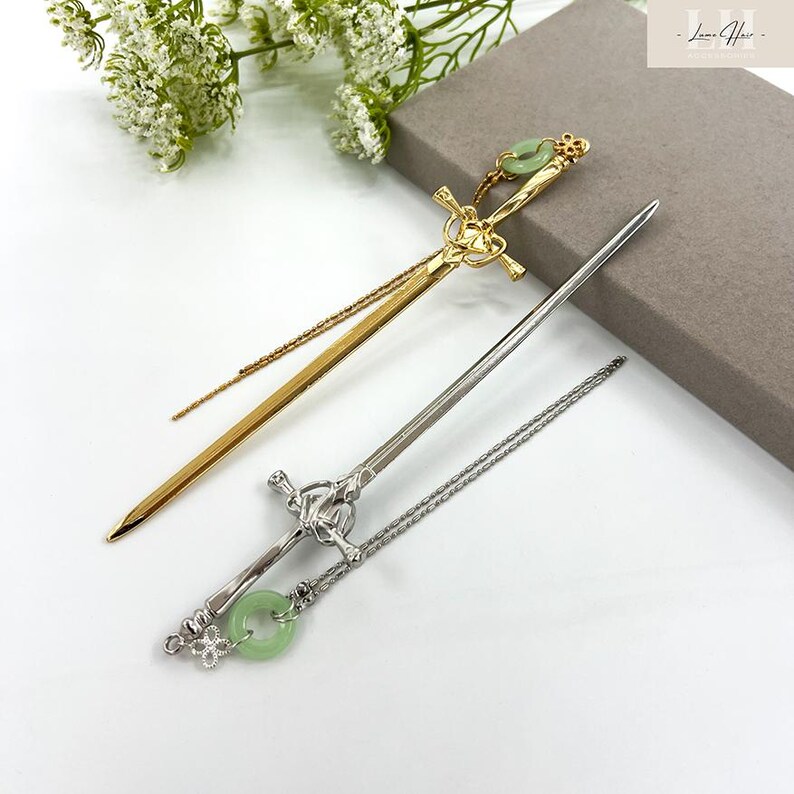 Op de afbeelding: Twee decoratieve haarstokjes. E&eacute;n is goudkleurig met een zwaardontwerp en een groene cirkelvormige accent. De andere is zilverkleurig met een zwaardontwerp en een groene cirkelvormige accent. Beide hebben ingewikkelde details en worden op een wit oppervlak weergegeven.