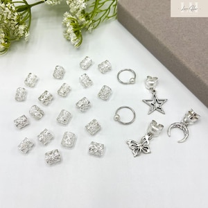 May include: A collection of silver hair accessories, including hair cuffs with star, butterfly, and crescent moon charms, along with small hoop earrings and decorative hair beads. The accessories are displayed on a white surface.