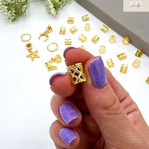 May include: A hand holding a gold-colored, open-weave hair bead. Various other gold hair accessories, including star, crescent moon, and ring-shaped beads, are scattered on a white surface. The person's nails are painted a sparkly purple.