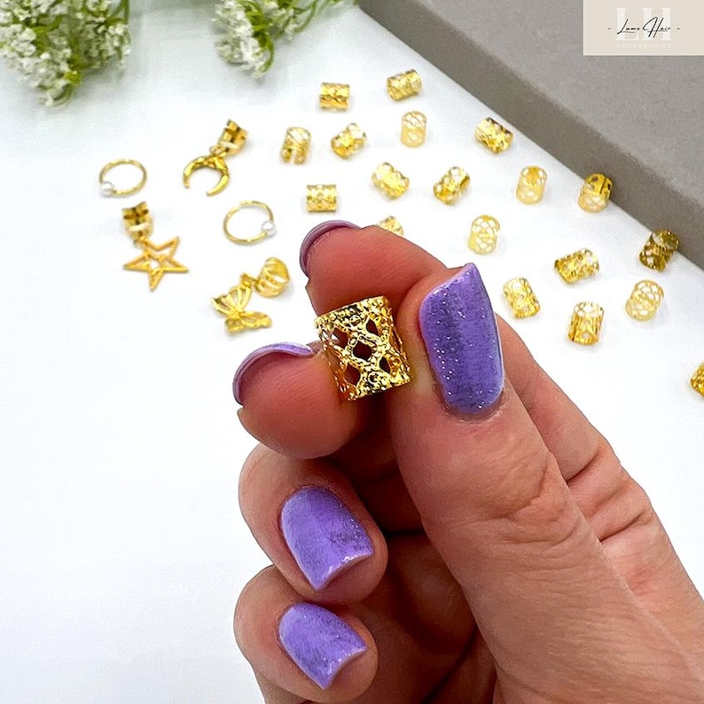 May include: A hand holding a gold-colored, open-weave hair bead. Various other gold hair accessories, including star, crescent moon, and ring-shaped beads, are scattered on a white surface. The person's nails are painted a sparkly purple.