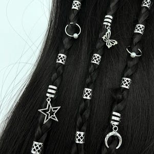 May include: Close-up of black braided hair adorned with silver hair beads and charms. Charms include a butterfly, star, crescent moon, and small rings. The beads have a geometric design. The hair accessories add a decorative touch to the hairstyle.