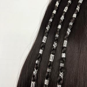 以下が含まれることがあります： 銀色のヘアビーズで飾られた3つの黒い三つ編みのクローズアップ。三つ編みは、長方形、円形、花の模様など、さまざまな銀色のビーズで装飾されています。髪は暗く滑らかで、明るい銀色のアクセントとコントラストをなしています。