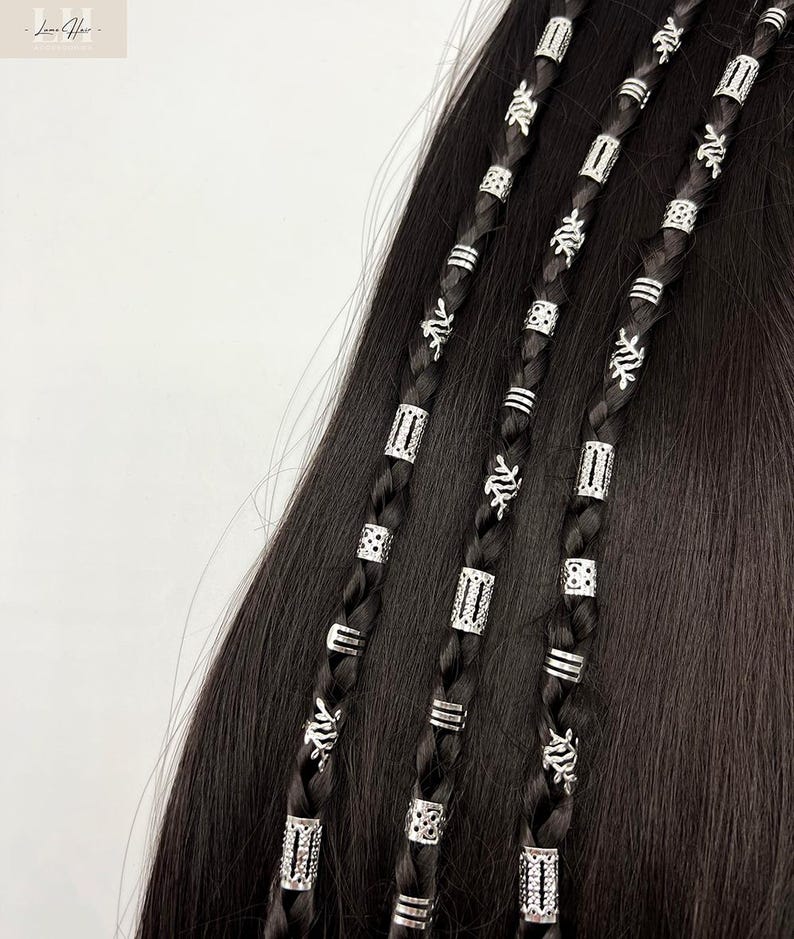 以下が含まれることがあります： 銀色のヘアビーズで飾られた3つの黒い三つ編みのクローズアップ。三つ編みは、長方形、円形、花の模様など、さまざまな銀色のビーズで装飾されています。髪は暗く滑らかで、明るい銀色のアクセントとコントラストをなしています。