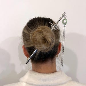 Op de afbeelding: Een zilveren haarspeld met een zwaardachtig ontwerp en een groene cirkelvormige bedel. Een tweede zilveren haarspeld is ook zichtbaar. De haaraccessoires zijn in een knot kapsel geplaatst. De afbeelding is tegen een witte achtergrond.