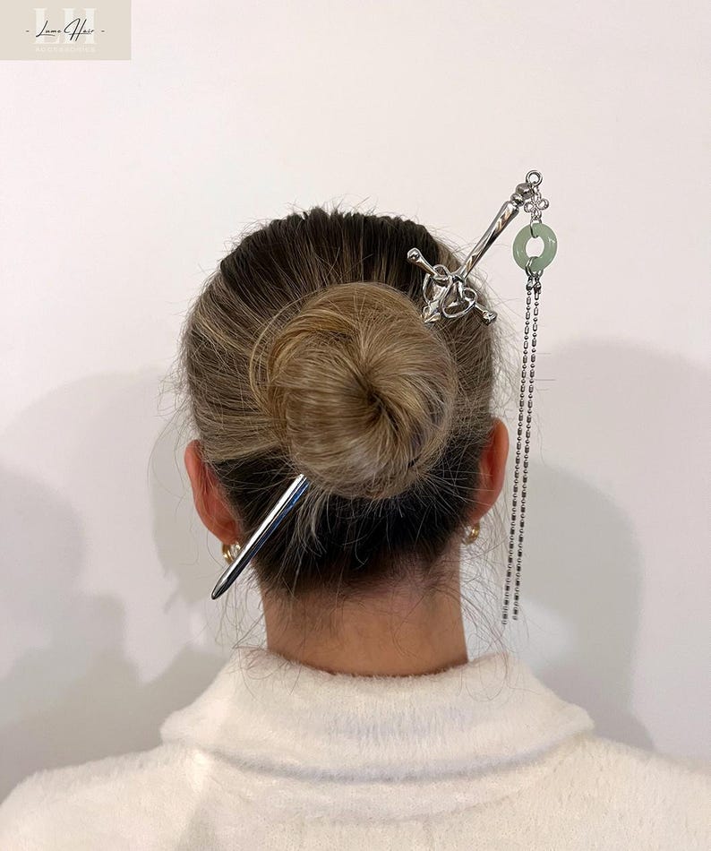 Op de afbeelding: Een zilveren haarspeld met een zwaardachtig ontwerp en een groene cirkelvormige bedel. Een tweede zilveren haarspeld is ook zichtbaar. De haaraccessoires zijn in een knot kapsel geplaatst. De afbeelding is tegen een witte achtergrond.