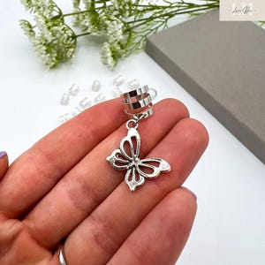 May include: Silver-toned ear cuff featuring a butterfly charm. The cuff has a triple-band design that wraps around the ear. The butterfly charm is intricately detailed with openwork wings. The jewelry is displayed against a soft background.
