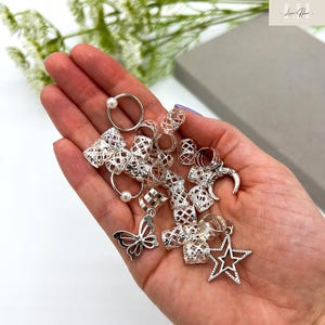 May include: A collection of silver hair accessories, including rings, charms, and decorative pieces, are displayed in an open hand. The accessories feature various designs, such as a butterfly, a star, and openwork patterns. Some pieces include small pearls.