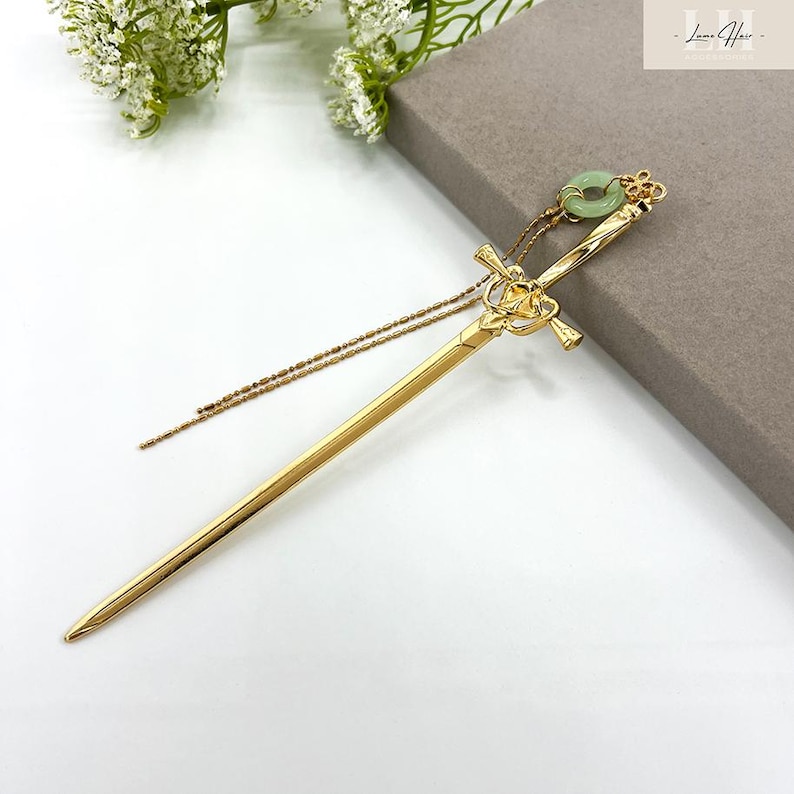 Op de afbeelding: Een goudkleurige haarspeld met een zwaardachtig ontwerp. De speld heeft een decoratief handvat met ingewikkelde details en een kleine, groene cirkelvormige steen aan de bovenkant. Een delicate gouden ketting hangt aan het handvat. Het haaraccessoire wordt op een wit oppervlak weergegeven.