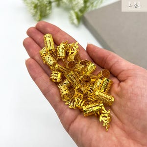 以下が含まれることがあります： 様々な円筒形をした金色のヘアビーズのコレクション。一部は複雑な透かし彫りのデザイン。手の中にビーズが収められ、金属的な輝きと精巧な職人技が際立っています。背景は柔らかいニュートラルカラーです。