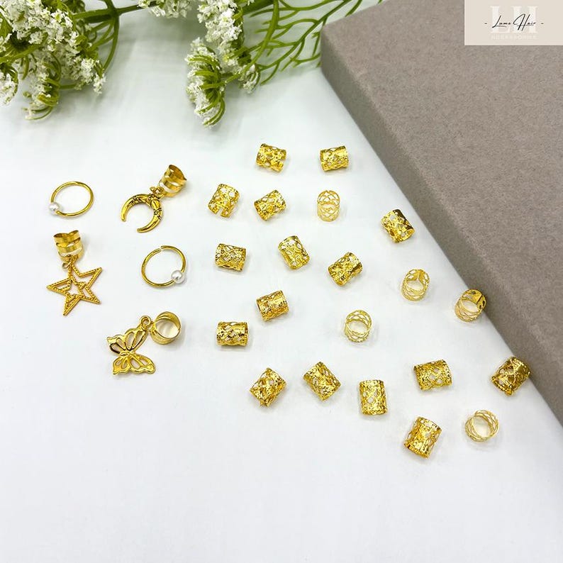 May include: A collection of gold hair accessories, including rings, charms with stars, moons, and butterflies, and decorative beads. The accessories are scattered on a white surface, with a gray rectangular object in the background.