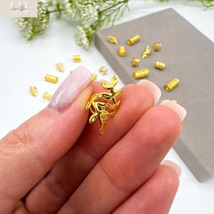 以下が含まれることがあります： 手に持たれた金色の葉の形をしたチャームのクローズアップ。周りには、さまざまな小さな金色のビーズやチャームが散らばっています。背景には灰色の表面と白い花が含まれています。
