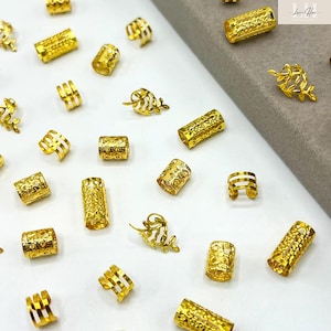 以下が含まれることがあります： 金色のヘアアクセサリーのコレクション。テクスチャ加工と透かし彫りのデザインの円筒形ビーズ、葉の形をしたアクセントが含まれています。白いストライプが入ったビーズもあります。白い表面に散らばっています。