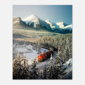 May include: A bright orange freight train travels along a curved track through a snowy landscape. The scene features dense, snow-covered evergreen trees in the foreground and a large, majestic mountain range with snow-capped peaks in the background.