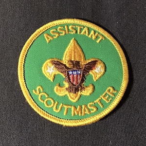 May include: A circular green embroidered patch with gold trim. The patch features the words "ASSISTANT" and "SCOUTMASTER" in gold lettering. The center design includes a gold fleur-de-lis and a brown eagle with a red, white, and blue shield.