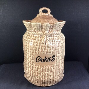 May include: Tan ceramic cookie jar with a woven basket design. The lid has a rope handle. The word "Cookies" is printed on a rectangular label on the front of the jar. The jar is a light brown color.