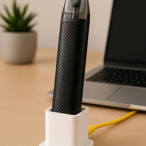 Może przedstawiać: Czarny e-papieros z teksturowanym uchwytem znajduje się w białej stacji ładującej. Urządzenie ma przezroczysty ustnik. Żółty kabel ładujący jest podłączony do stacji dokującej. Laptop i mała roślina doniczkowa znajdują się w tle.