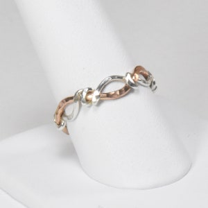 May include: A silver and copper ring with a twisted, interwoven design. The ring is on a white display stand.