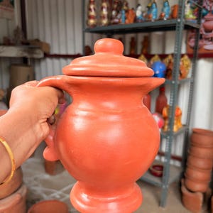 Op de afbeelding: Een terracotta kleikruik met een rond lichaam, een brede hals, een handvat aan de zijkant en een bijpassend deksel. Het vat heeft een gladde, matte, rood-oranje afwerking en staat op een voetstuk, met een traditioneel handgemaakt ontwerp.
