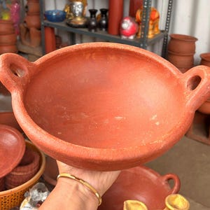 May include: A round, terracotta-colored clay bowl with a wide, open top and two integrated side handles. The bowl features a matte, earthy finish with a slightly textured surface, showcasing a simple, functional design for holding various items.