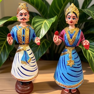 May include: Two hand-painted figurines depicting a person in a blue and white outfit with a turban and a person in a blue and pink outfit with a gold headpiece. Both figures have raised hands with red diamond markings and stand on brown circular bases.