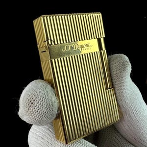 May include: A rectangular gold-tone metal lighter featuring a vertical ribbed texture across the body. The front surface displays the engraved text S.T. Dupont Paris on a smooth horizontal band that separates the top lid from the main body.