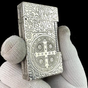 May include: A rectangular silver-toned metal lighter featuring an intricate engraved pattern and a central circular emblem containing a cross with the letters C S P B and the text N D S M D. The side of the lighter displays the engraved text FIAT L S M D.