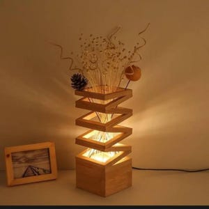 Könnte beinhalten: Eine Lampe aus hellem Holz mit quadratischem Sockel und einer spiralförmigen, offenen Rahmenstruktur, die ein warmes Licht ausstrahlt. Oben befindet sich ein dekoratives Arrangement aus getrockneten beigen Gräsern, kleinen weißen Blüten und einer runden braunen Samenkapsel.